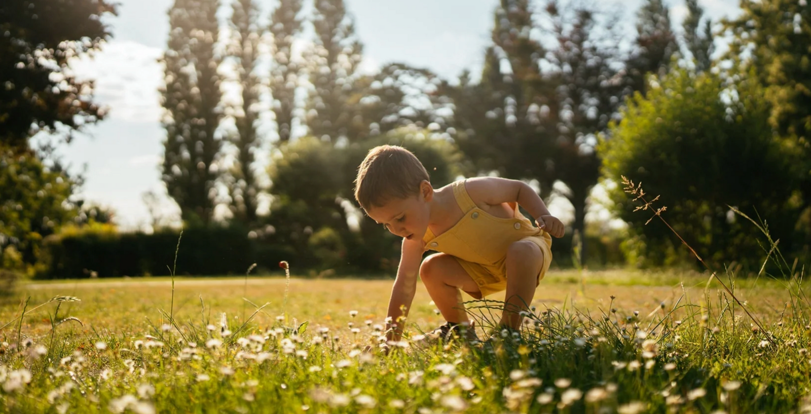 Toddler wearing a yellow outfit crouching in sunlit grass.