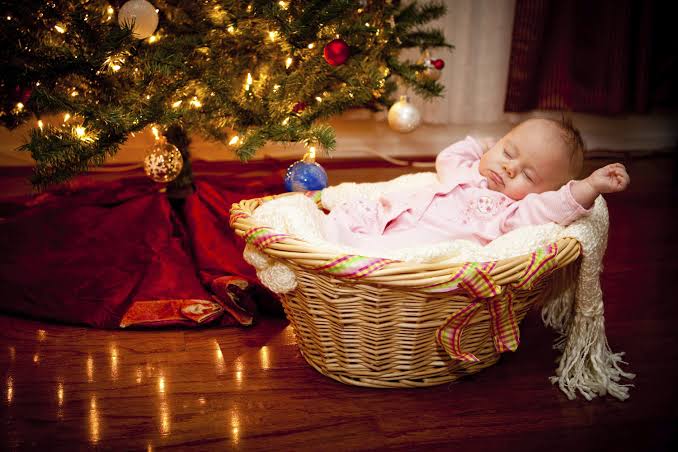 Baby in a pink outfit resting in a wicker basket beside a decorated Christmas tree